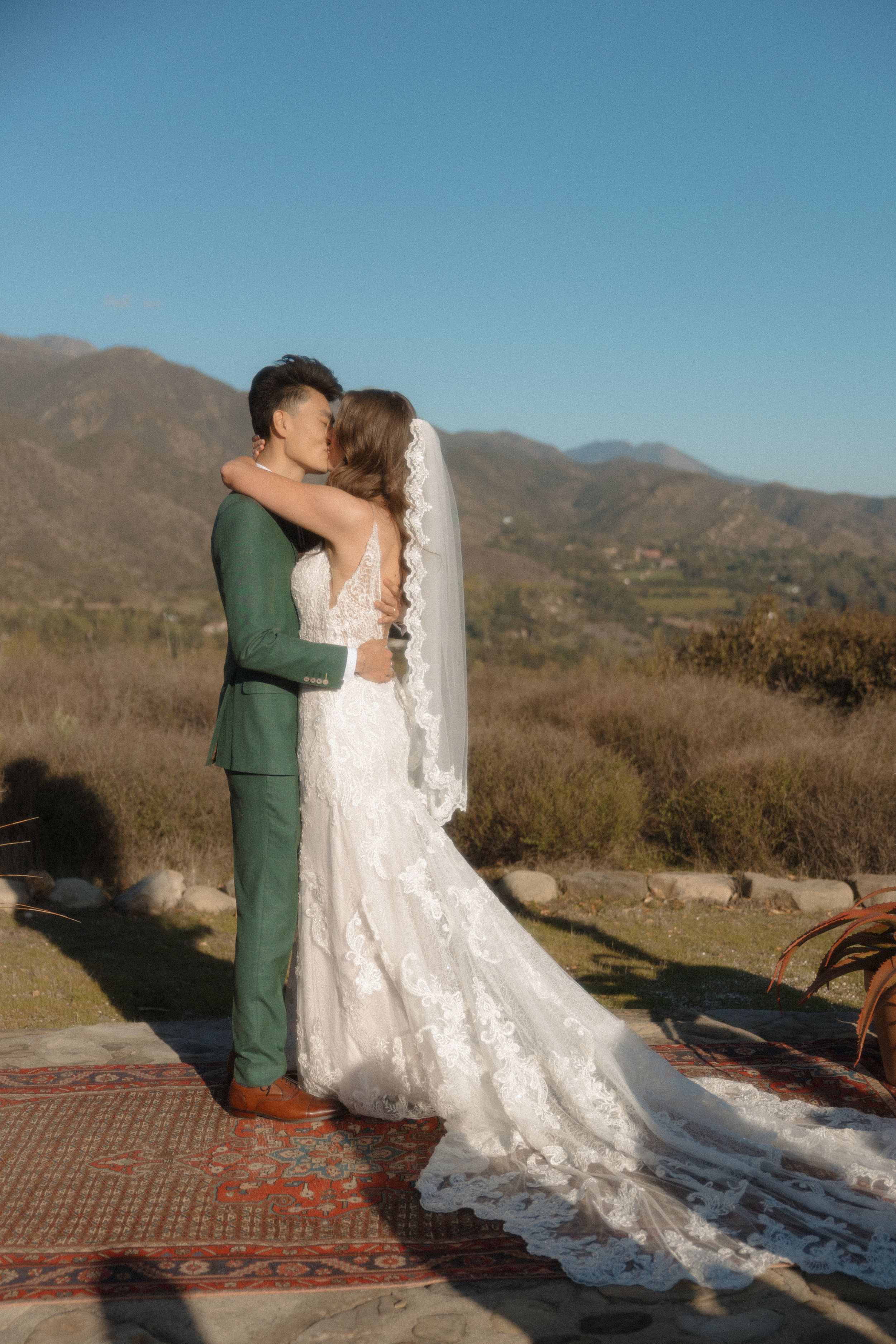 Newlyweds share first kiss in green suit and lace wedding gown with veil, scenic mountain landscape behind.