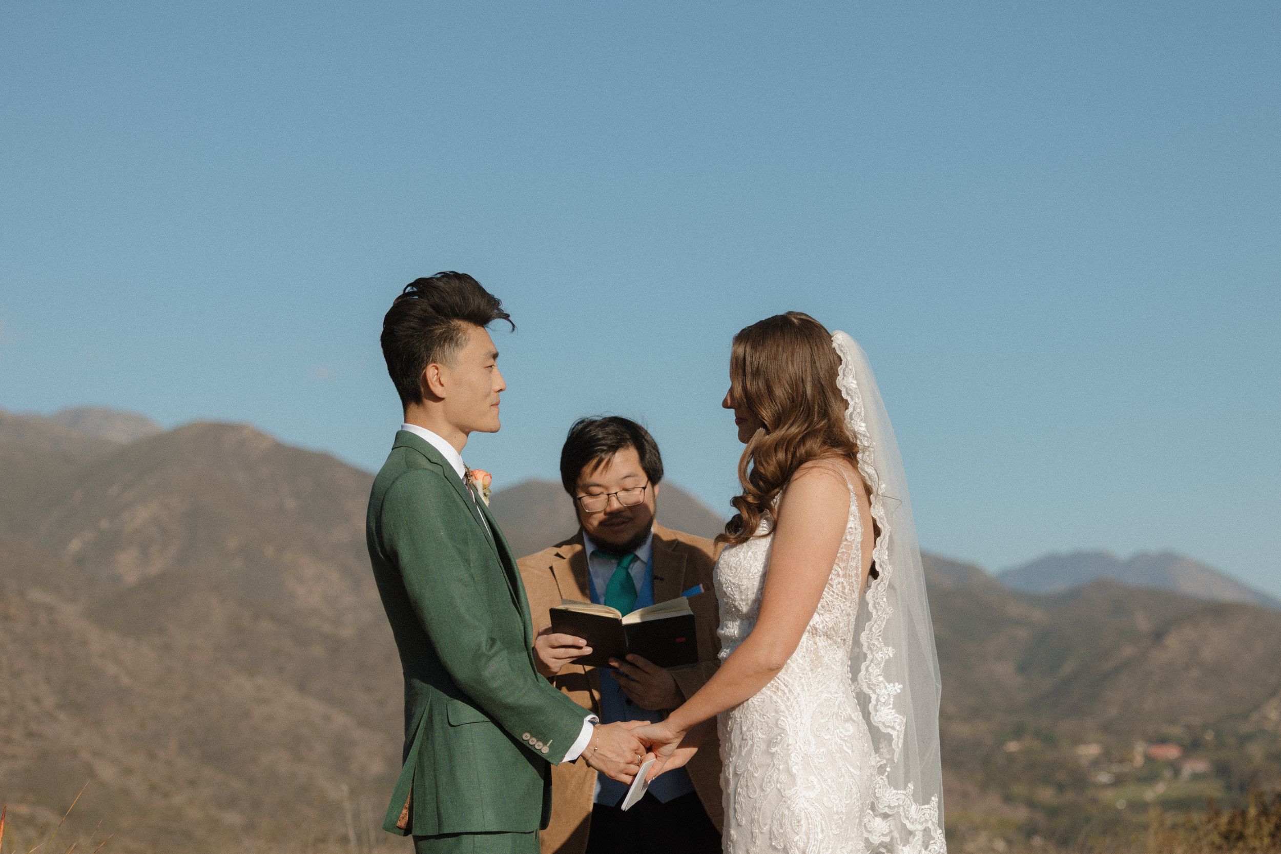 Outdoor wedding ceremony with couple exchanging vows before officiant, mountain backdrop and clear blue sky.