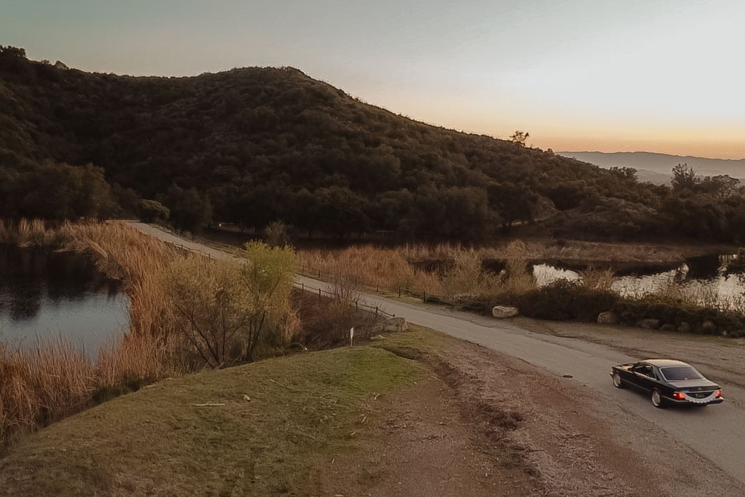 Aerial view of wedding car driving along scenic road beside lake at sunset.