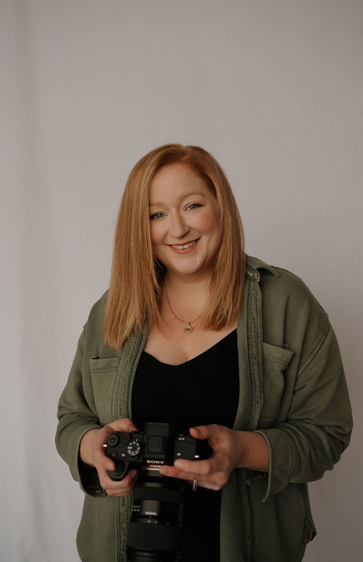 A smiling redhead woman sits relaxed on a black chair against a white fabric backdrop wearing a sage top and black pants.