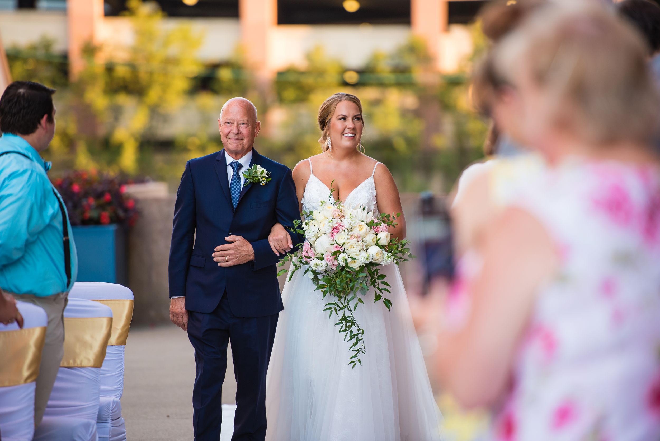 Bride walks down the aisle with her father at an outdoor wedding ceremony, holding a lush white and pink bouquet.
