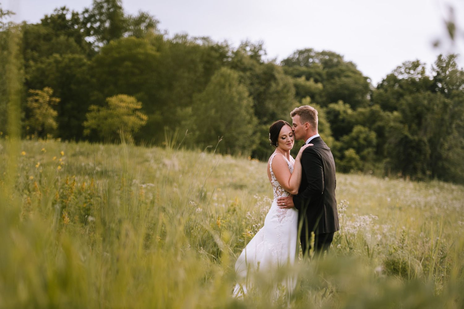 A bride and groom embrace in a lush green meadow, sharing a romantic moment on their wedding day.