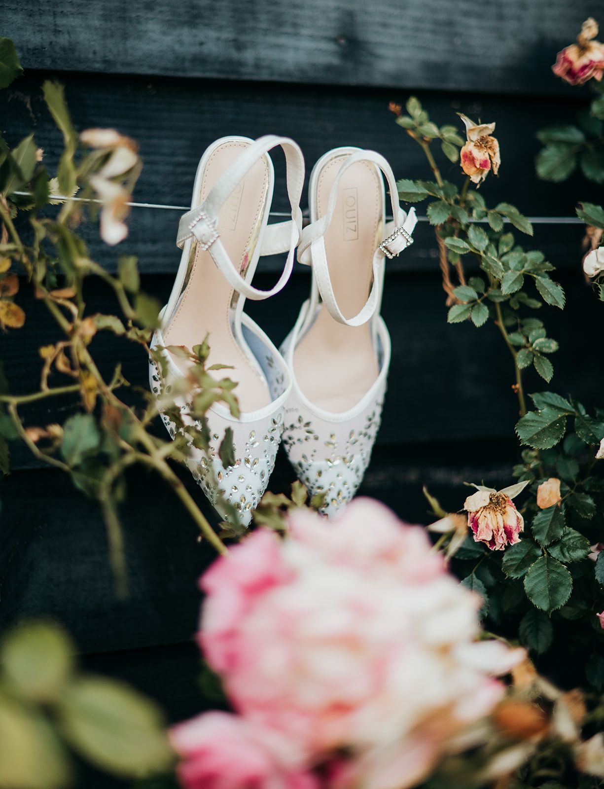 White embellished bridal heels with ankle straps displayed among pink roses and green foliage outdoors.