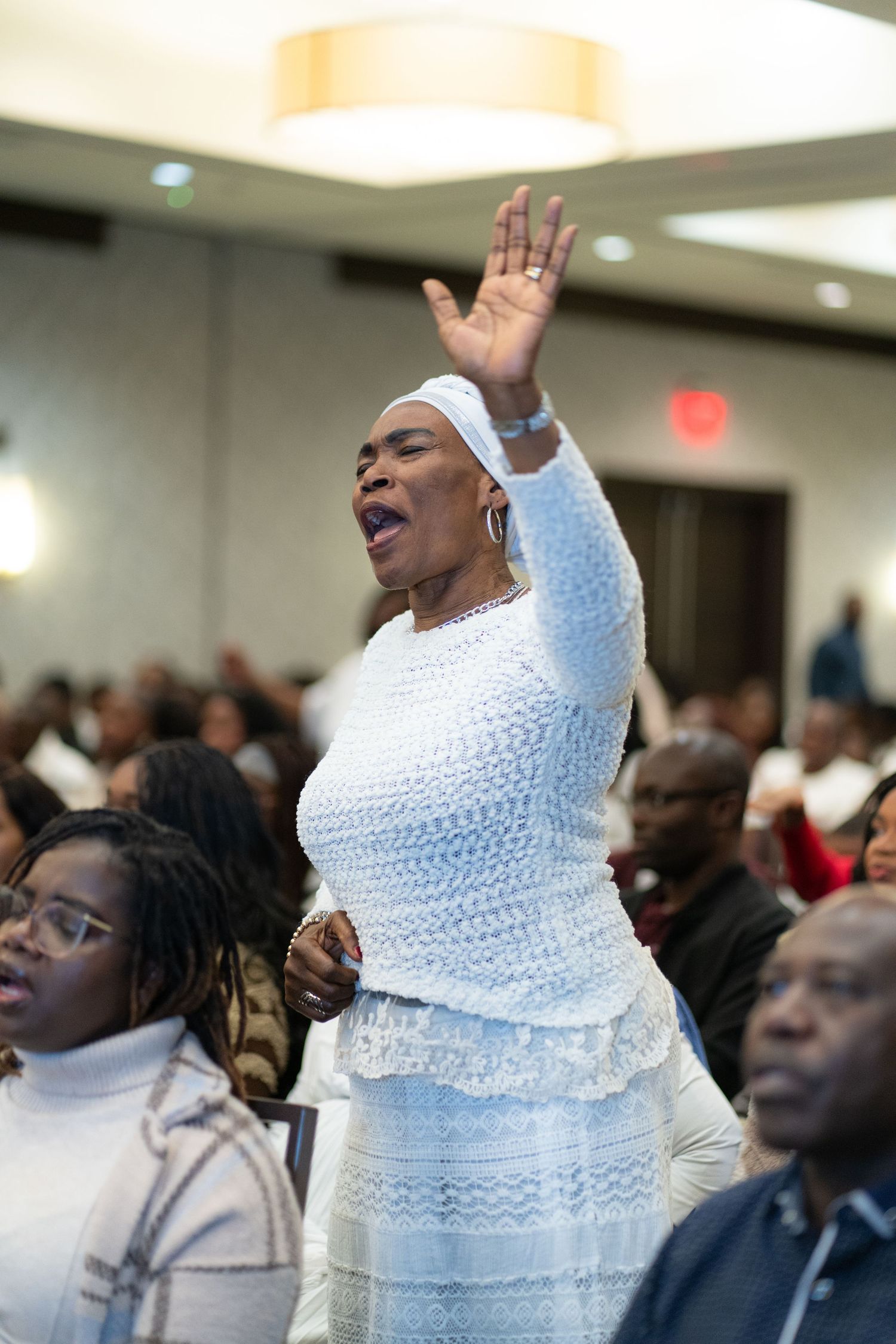 A person in white lace dress raises their hand and sings passionately during a church service or gathering.