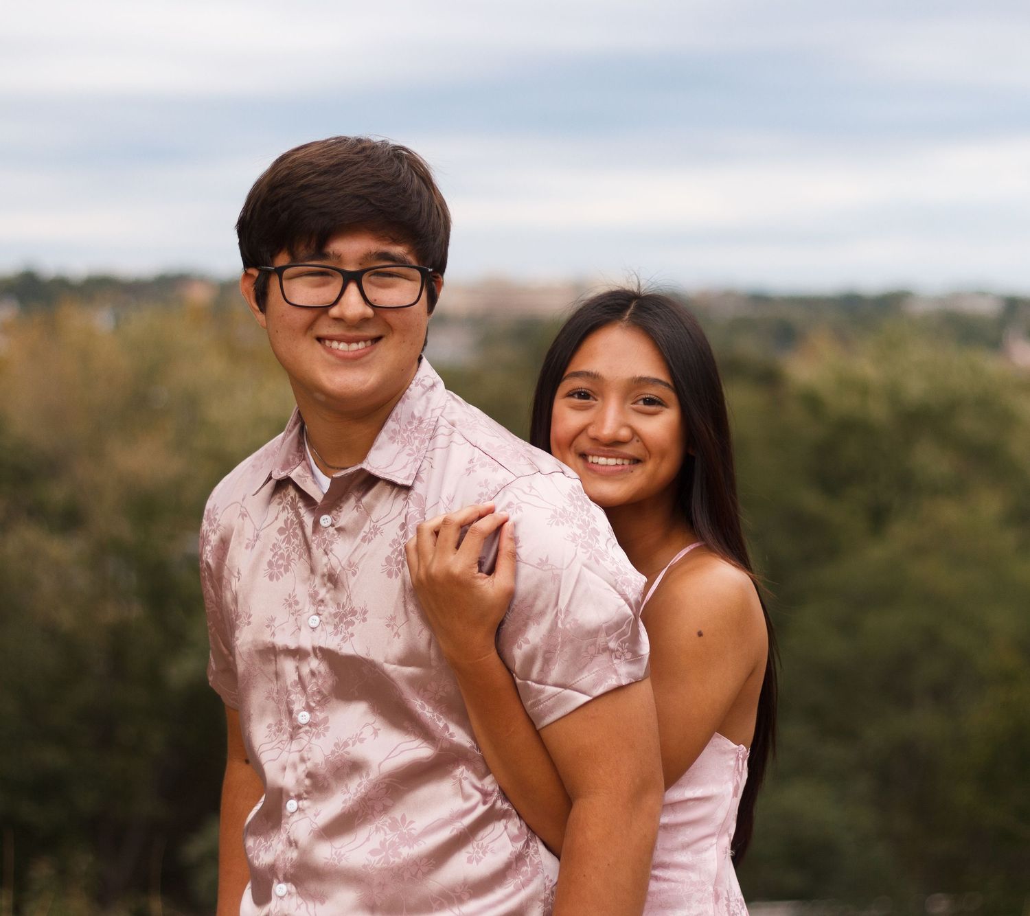 A joyful couple embraces outdoors on a cloudy day with trees and greenery in the background.