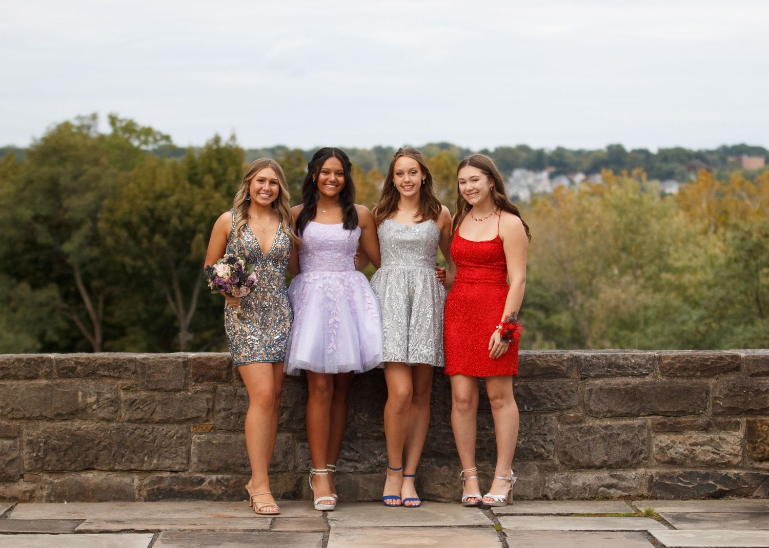 Four friends in sparkling cocktail dresses pose together on a stone wall with trees and autumn scenery behind them.