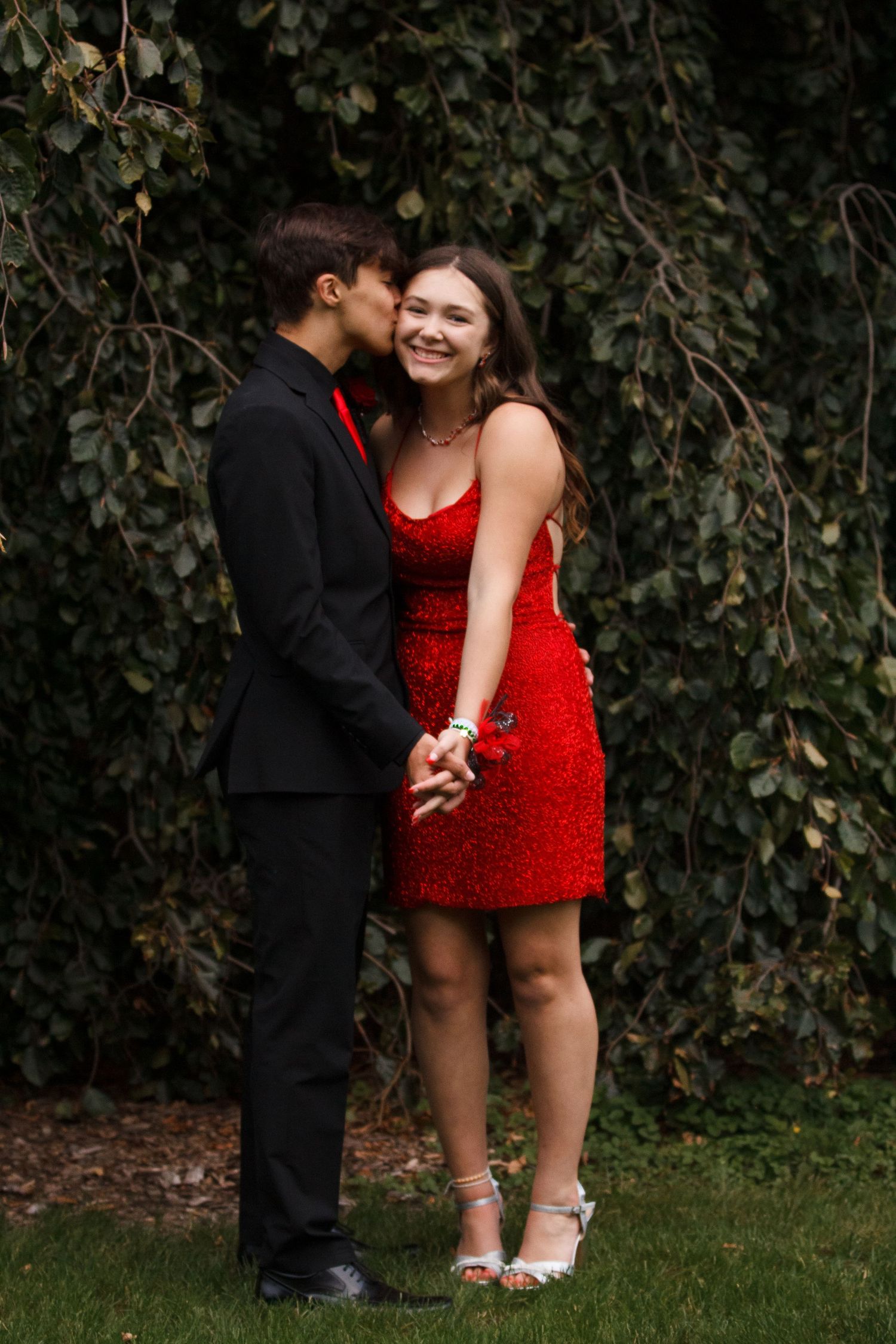 A couple shares a romantic moment outdoors, with one person in a black suit and another in a sparkly red cocktail dress.