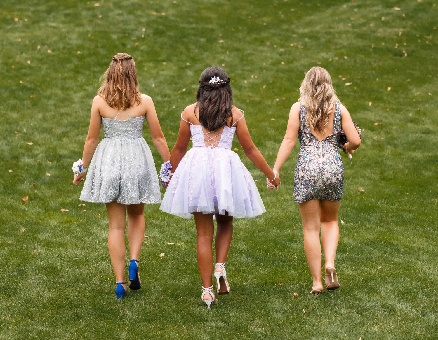 Three friends in short party dresses walk hand in hand across a green lawn on a sunny day.