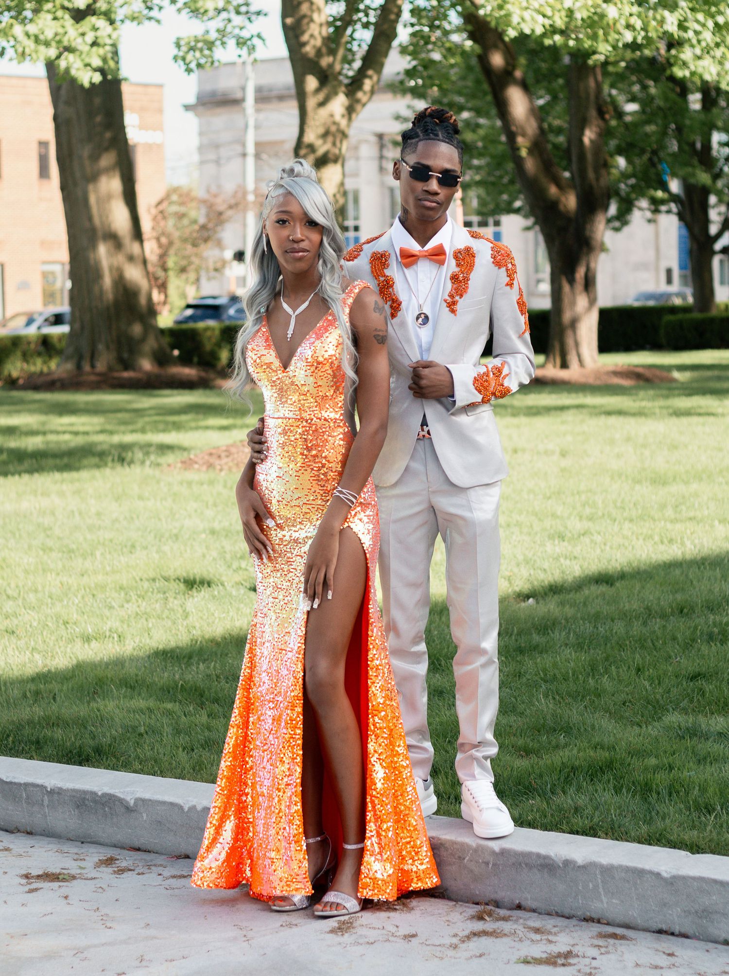 A couple in orange and white formal attire posing together outdoors on a sunny day in front of trees and buildings.