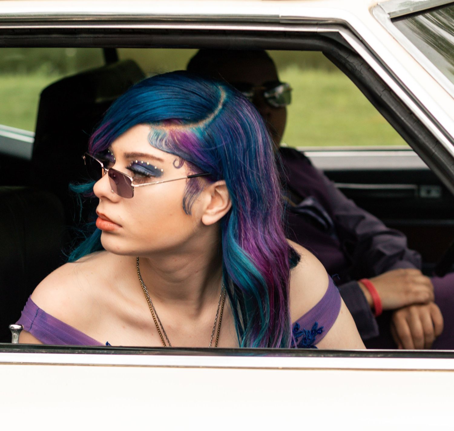 Person with vibrant blue and purple hair wearing sunglasses leans out car window on sunny day.Summer photo sessions Ohio, fall photos in You