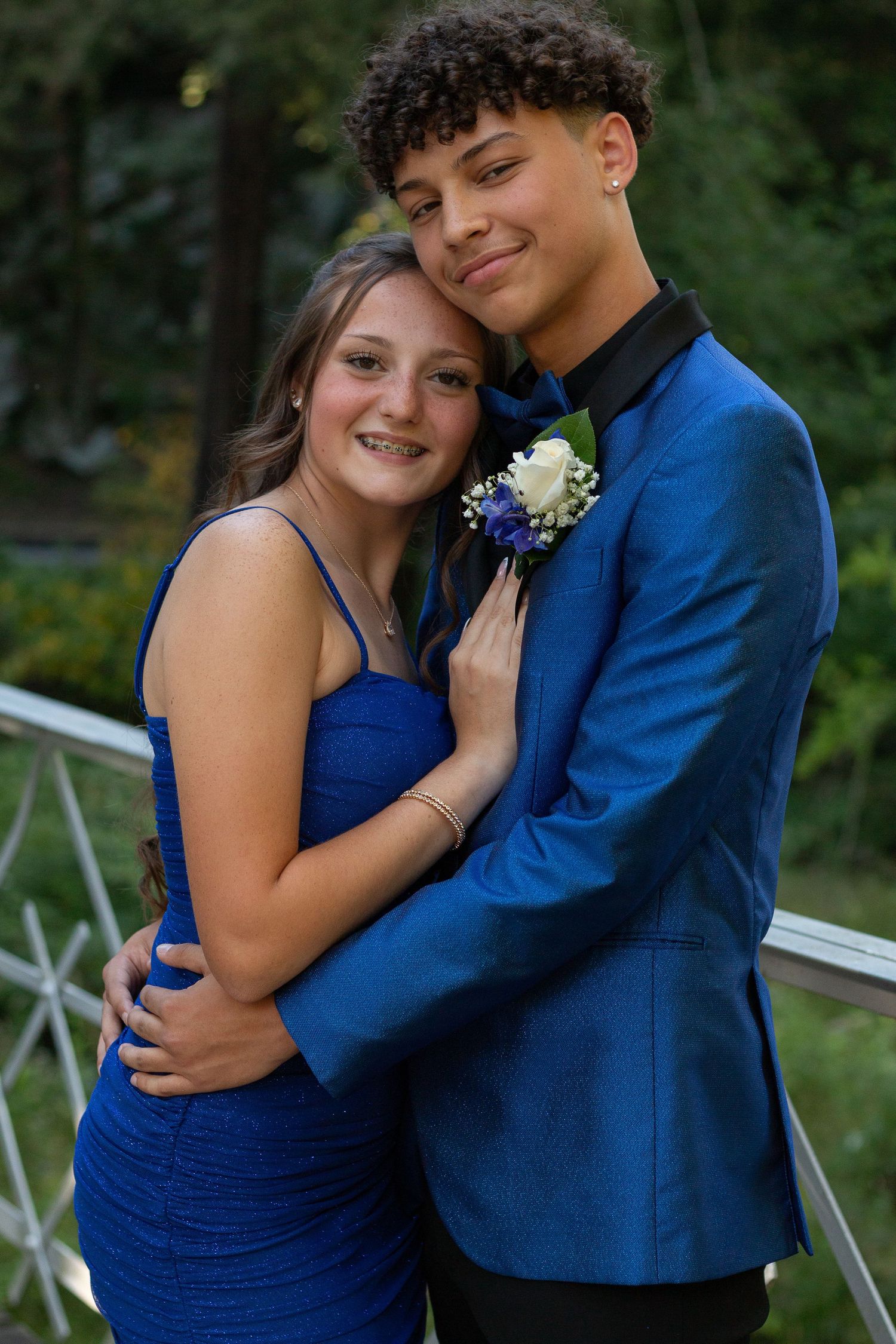 A couple in formal blue attire poses together on a white railing during what appears to be a prom or formal event.