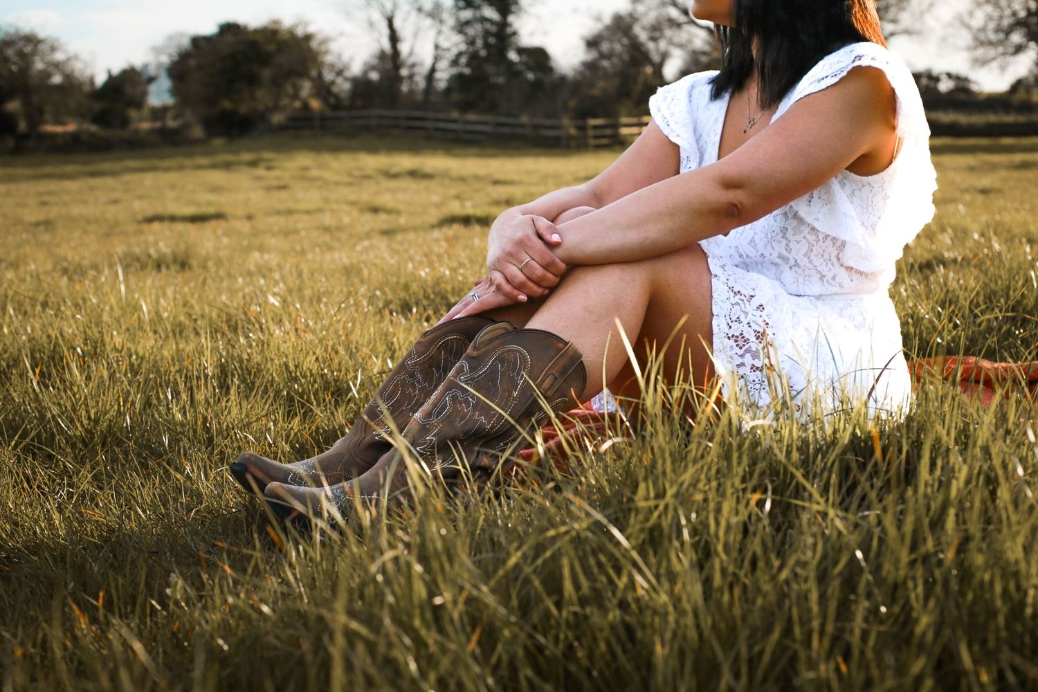 Person in white dress and brown boots sitting in sunlit grass field.