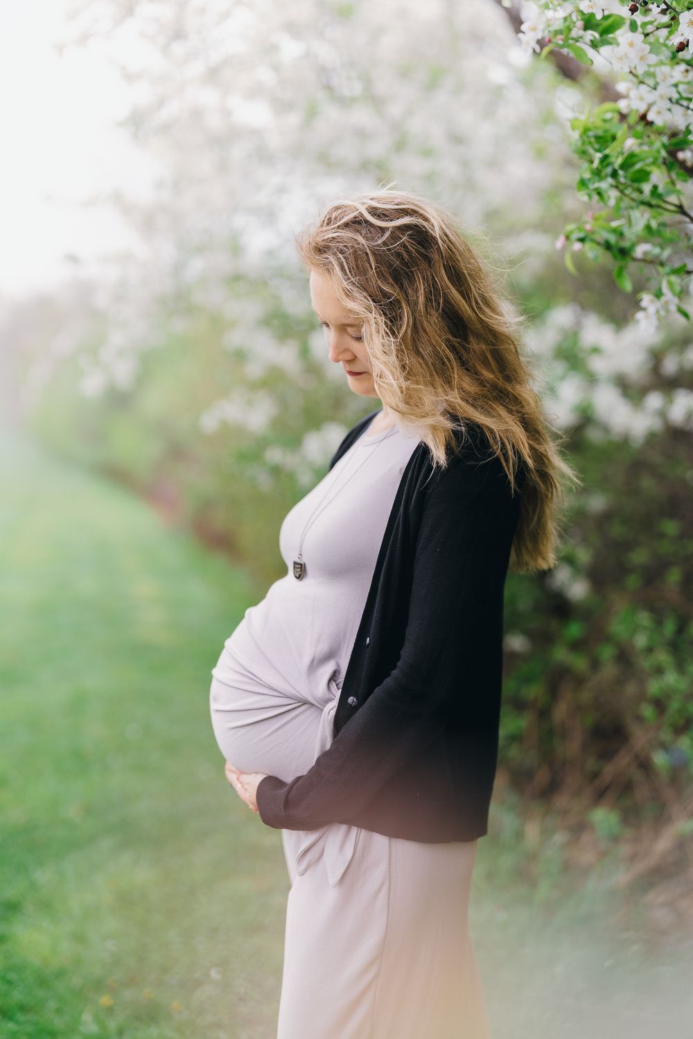 Expectant mother in white dress and black cardigan poses in front of spring blossoms in a garden setting at Barlow Road Farm Park in Ohio