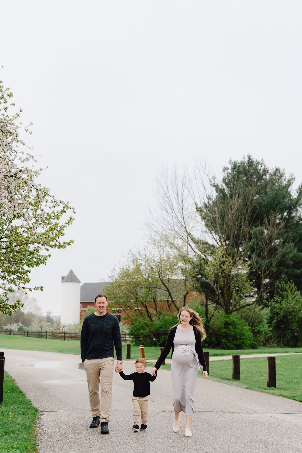 A family of three walking together on a paved park path surrounded by spring greenery and trees at Barlow Road Farm Park in Hudson, Ohio.