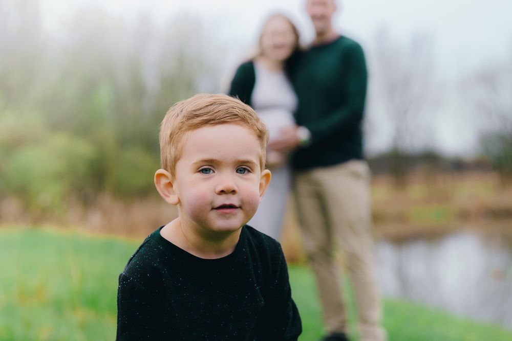 Young child smiles at the camera while standing outdoors with two people visible in the background at Wood Hollow Metro Park in Hudson, Ohio