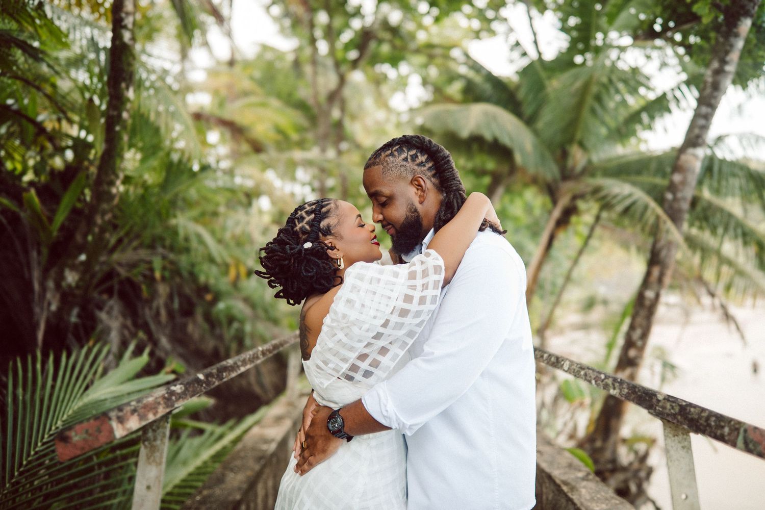 A loving couple embraces intimately under palm trees on a sunny tropical beach beside clear turquoise waters.