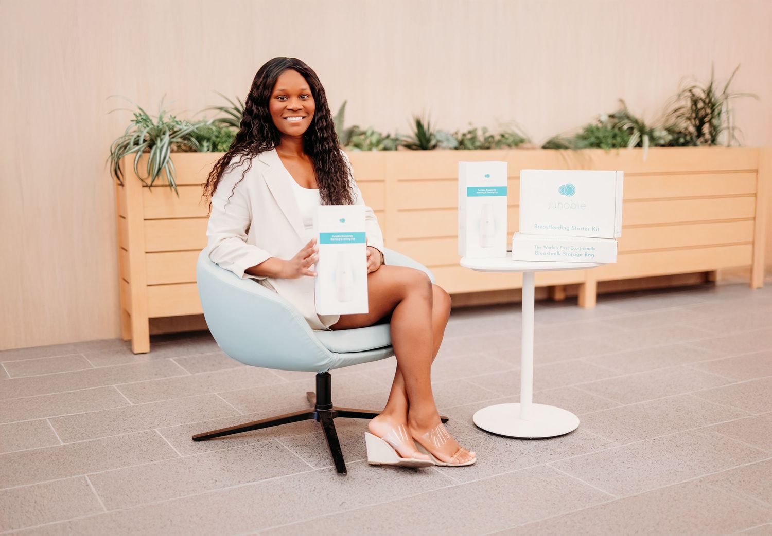 A professional wearing white blouse and light blue skirt sits in a modern chair with display stand and planter in background.