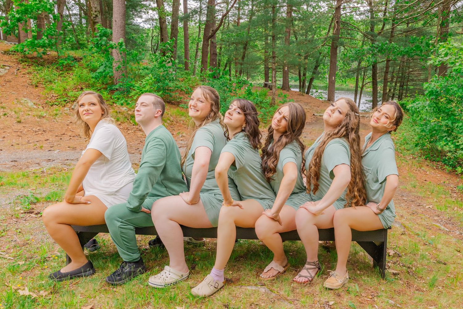 A group of friends sits casually in a line on an outdoor embankment surrounded by green forest scenery.