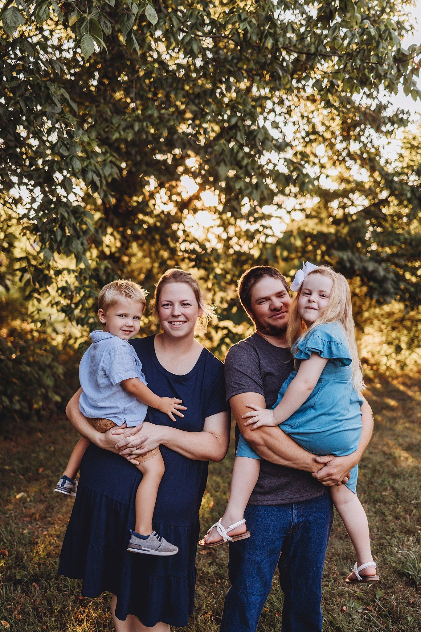 Family poses together outdoors during golden hour sunset with trees and sunlight filtering through leaves in background.