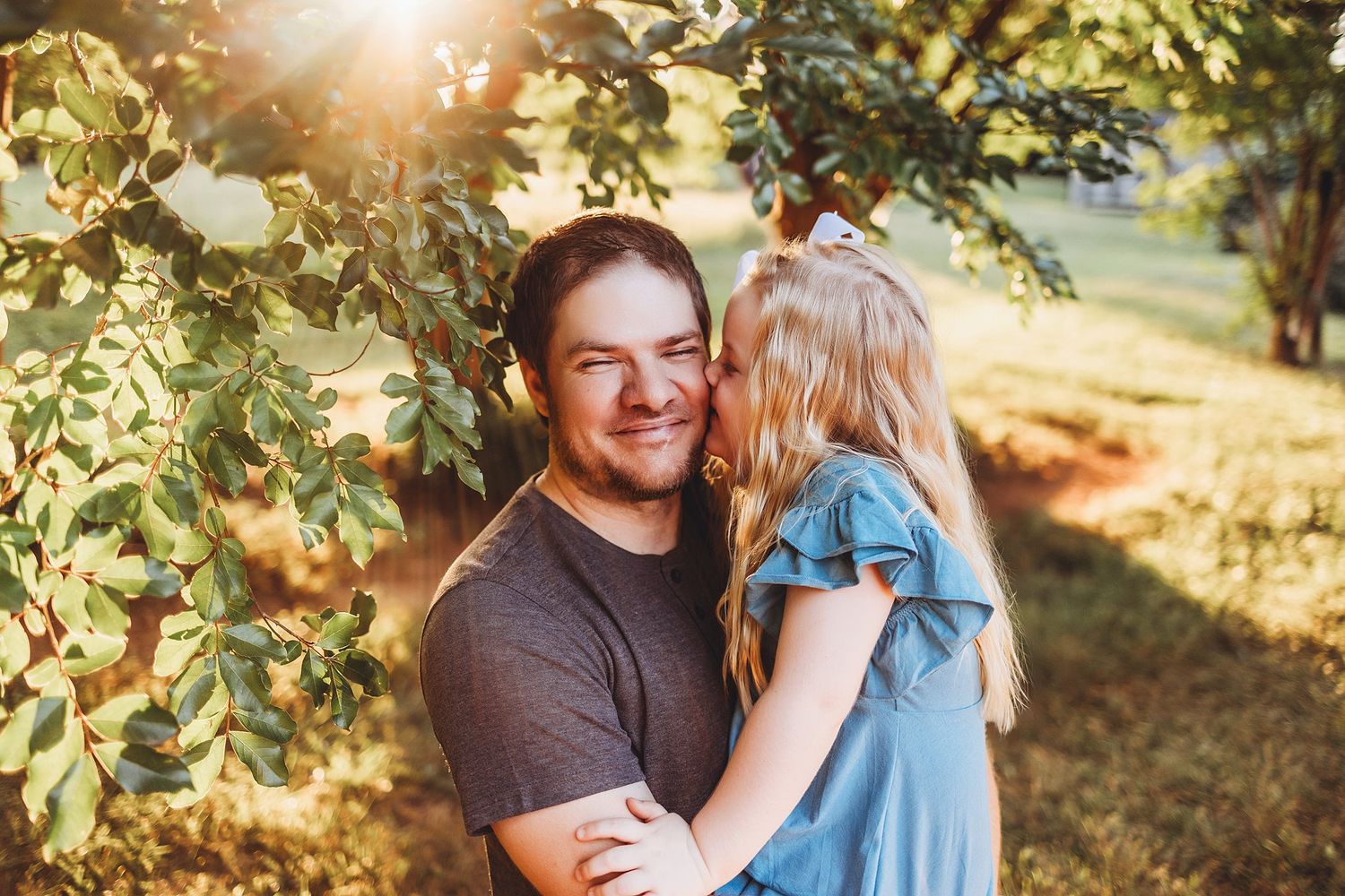 A candid moment captured in golden sunlight showing a warm embrace under tree branches at sunset.