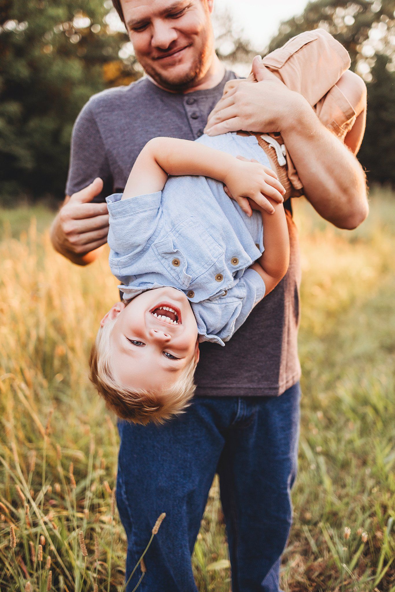 Father holding and playing with child upside down in golden field during sunset.