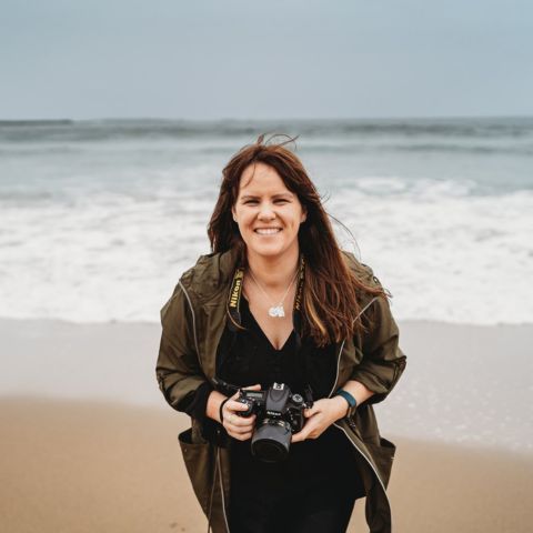 A photographer holding a vintage camera stands smiling on a cloudy beach with waves in the background.