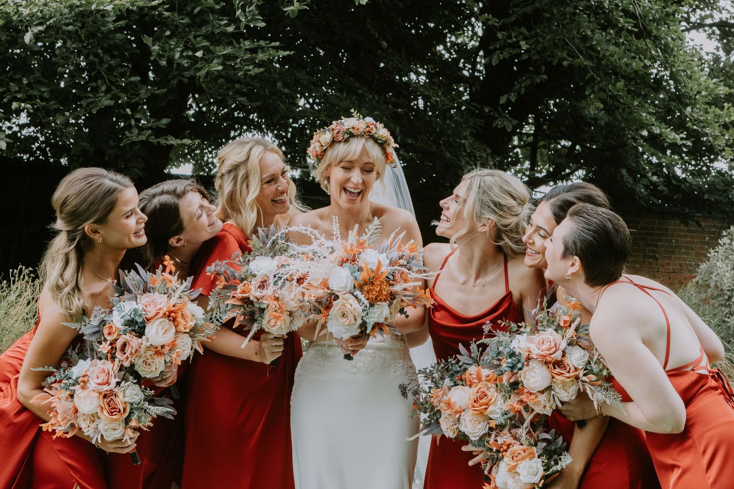 Wedding party in red bridesmaid dresses laughing joyfully while holding peach and ivory floral bouquets outdoors.