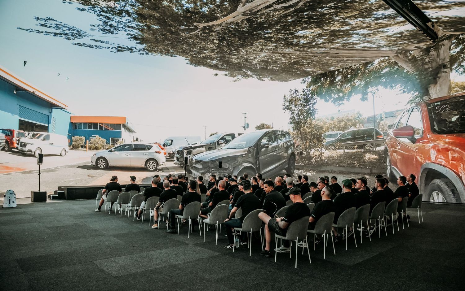 A large group in black attire seated on chairs underneath a concrete underpass with urban art and parked cars visible.