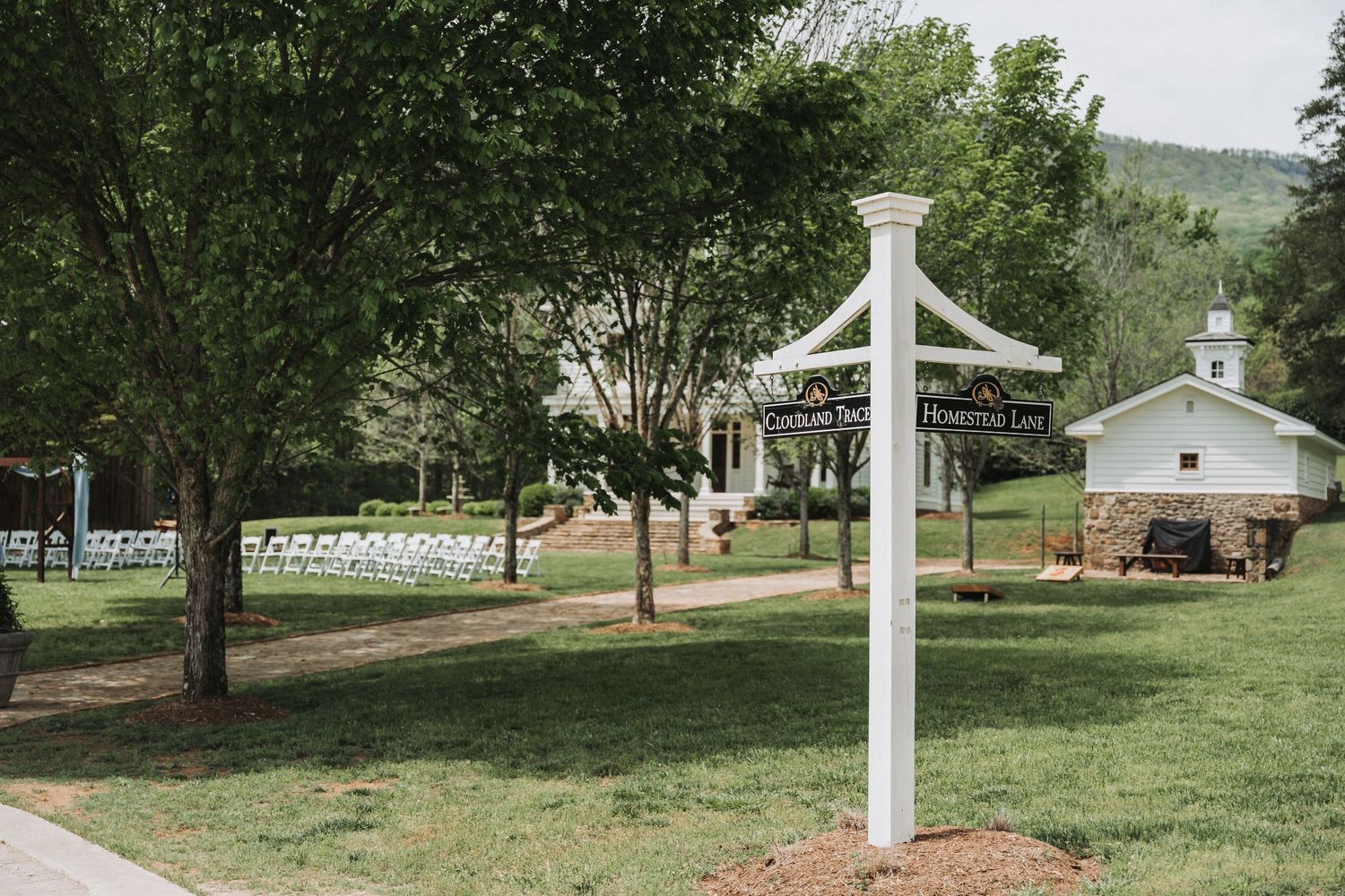 Outdoor wedding venue with a white cross and rows of white chairs on a grassy lawn surrounded by trees.