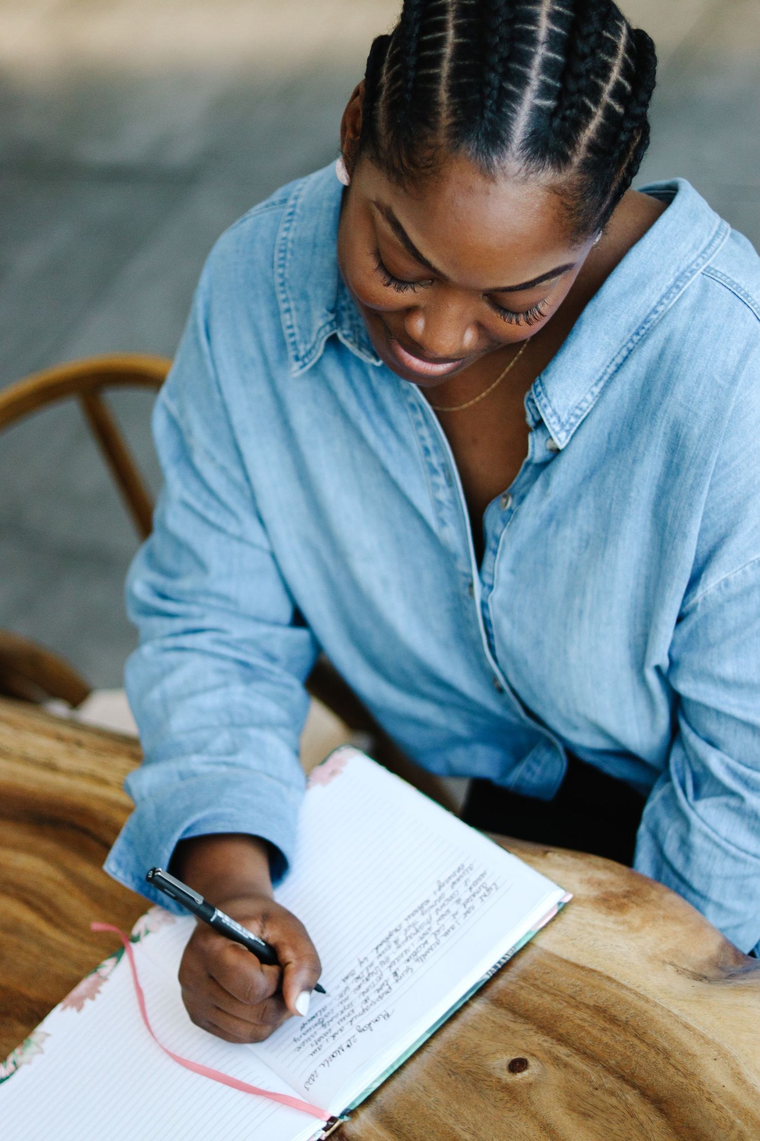 Person in denim shirt writing in notebook at wooden table.