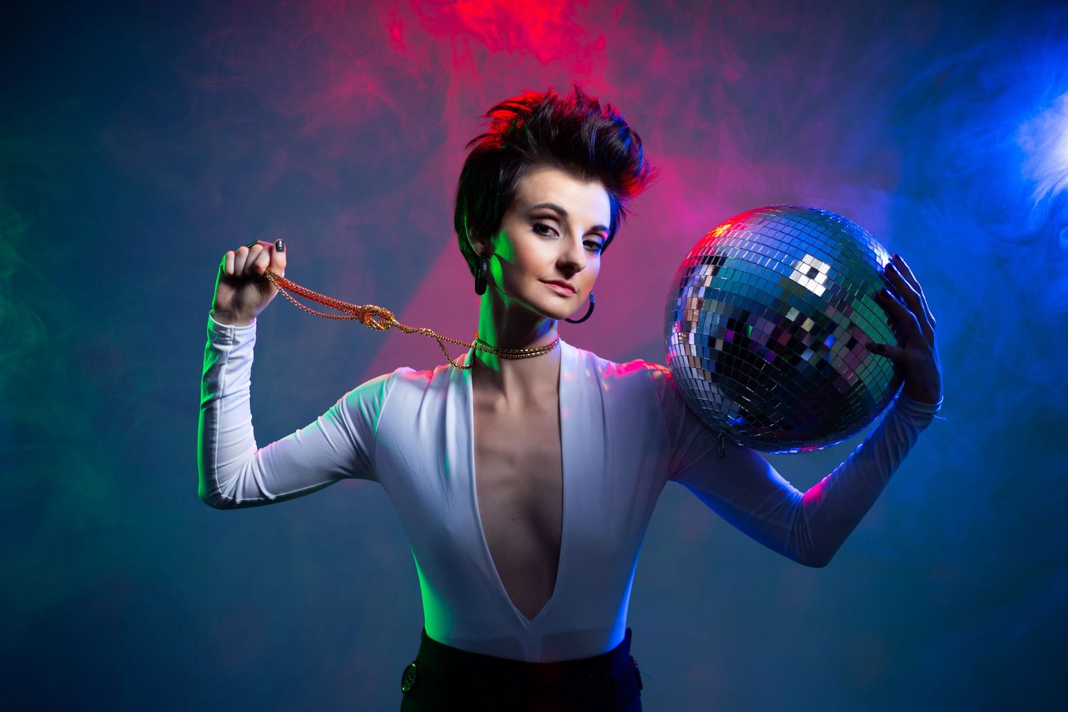 A stylishly dressed performer holds a disco ball against colorful red and blue smoke background in a dramatic studio shot.
