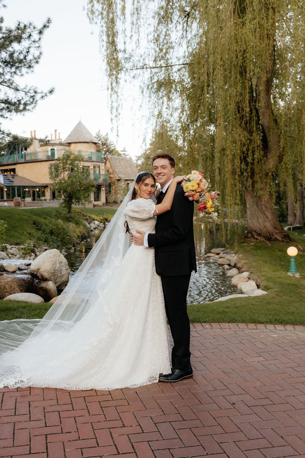Happy bride and groom posing together near a serene pond with weeping willow trees at their wedding venue.