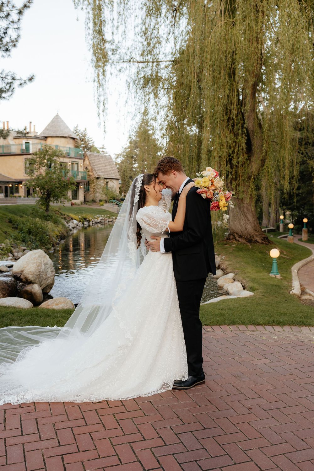 A bride and groom share a romantic kiss outdoors near a pond, surrounded by weeping willow trees.