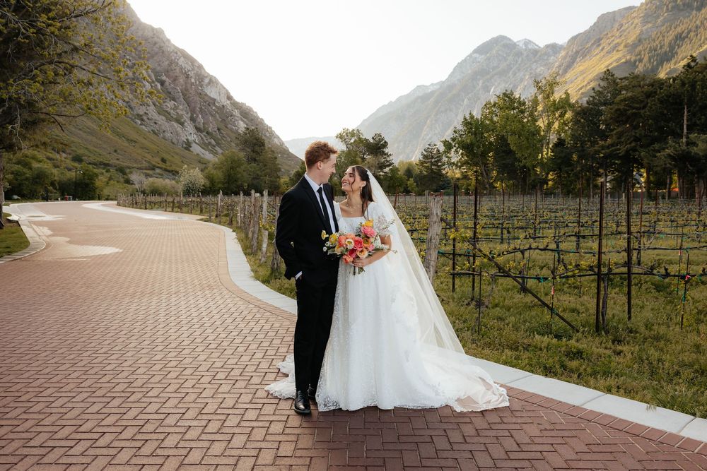 A bride and groom walk along a brick path through a vineyard with stunning mountain scenery behind them.