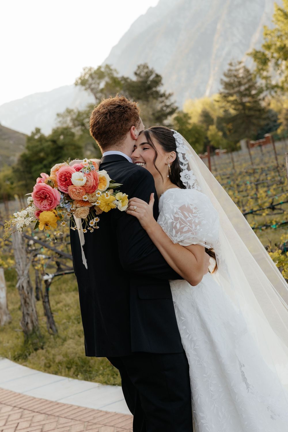 A joyful bride laughs as she embraces her groom in a vineyard setting with mountains in the background.