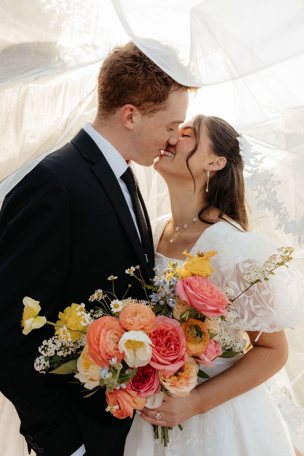 Bride and groom sharing an intimate kiss under a flowing veil while holding a colorful spring bouquet.