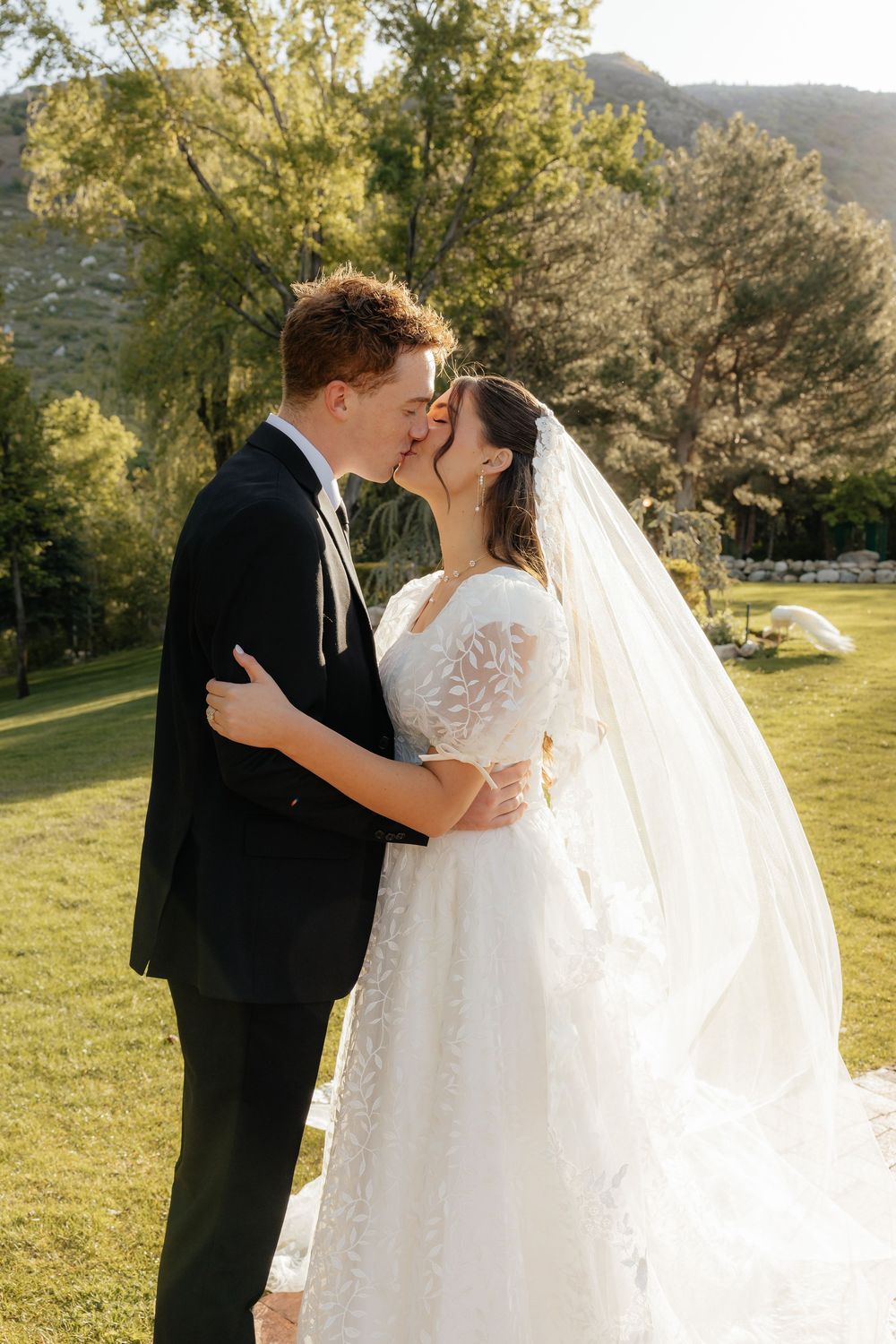 Bride and groom share a kiss on green lawn with mountain and swan visible in background.