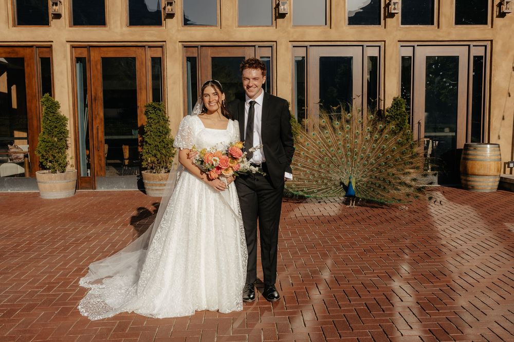 Bride and groom pose together on brick patio outside elegant venue with peacock in background.