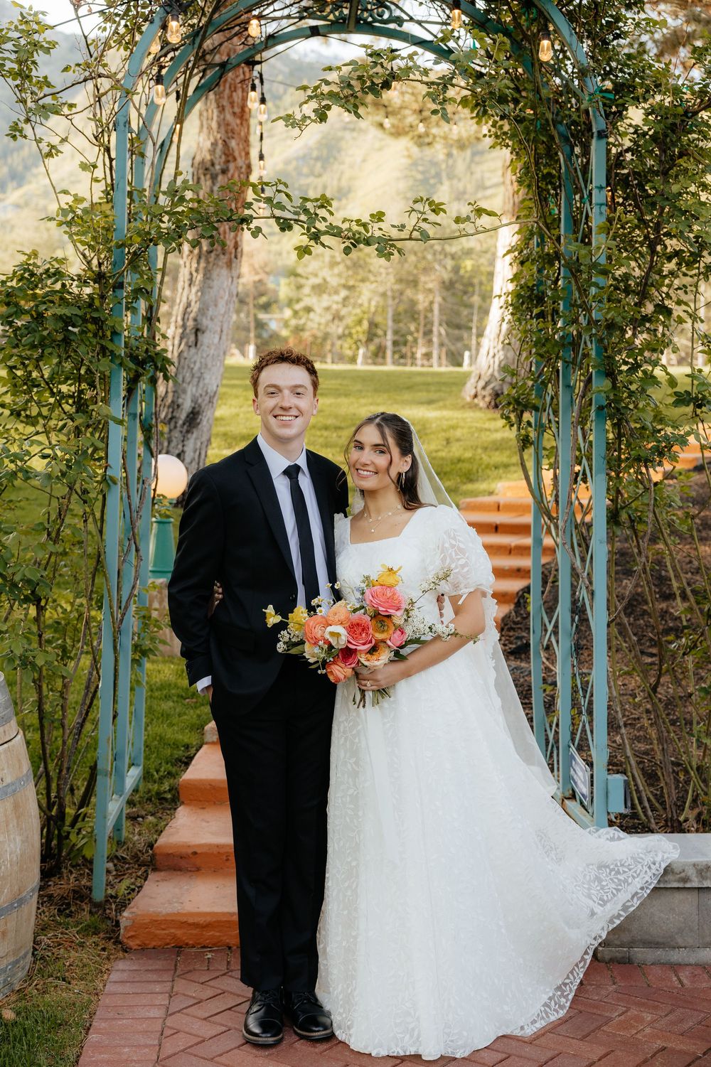 Smiling bride and groom stand together under teal garden archway with mountain backdrop.