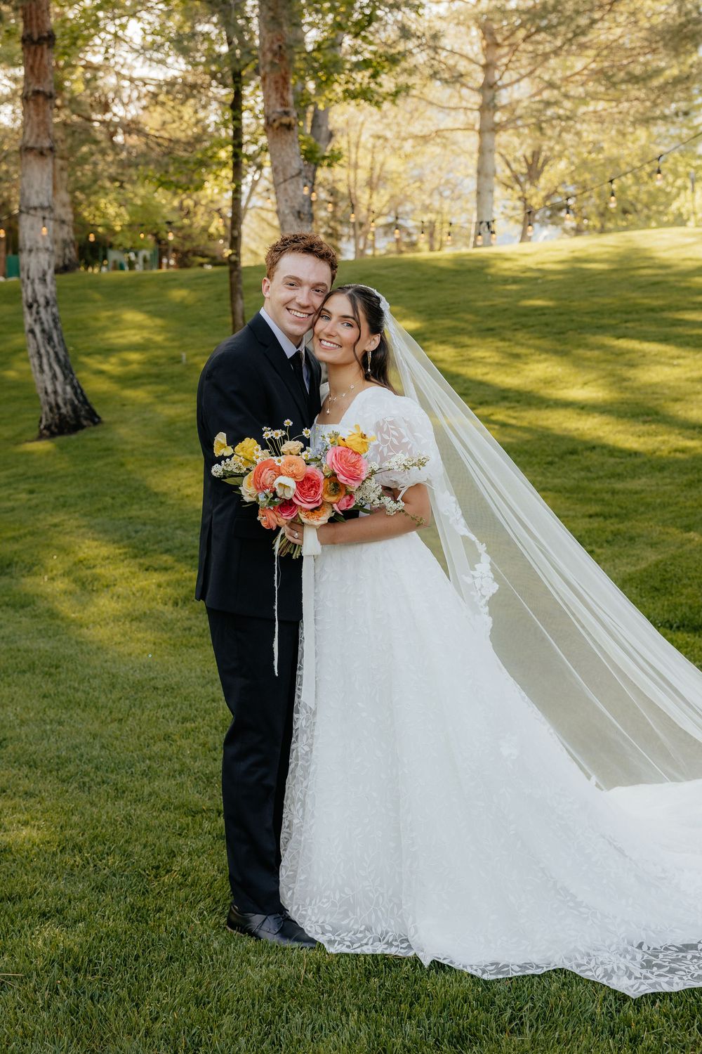 Bride and groom embrace on sunlit green lawn, bride holding vibrant coral and pink bouquet.