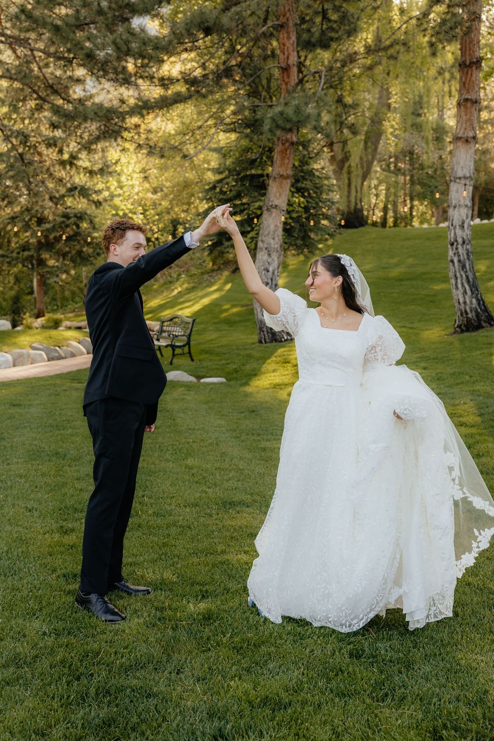 Joyful bride and groom dancing and spinning on a lush green lawn surrounded by tall pine trees.