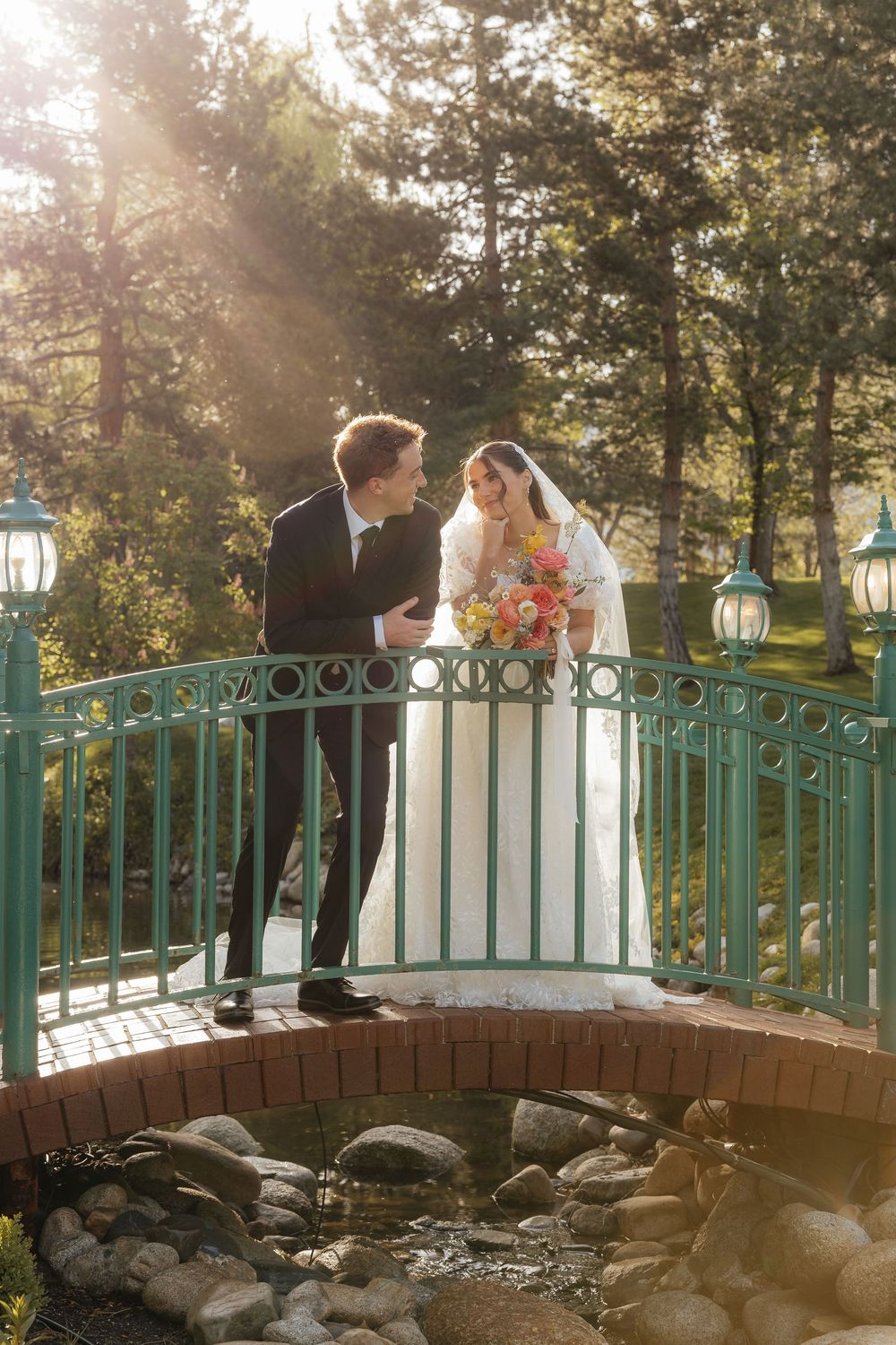 Bride and groom sharing a tender moment on a charming green bridge surrounded by sunlit pine trees.