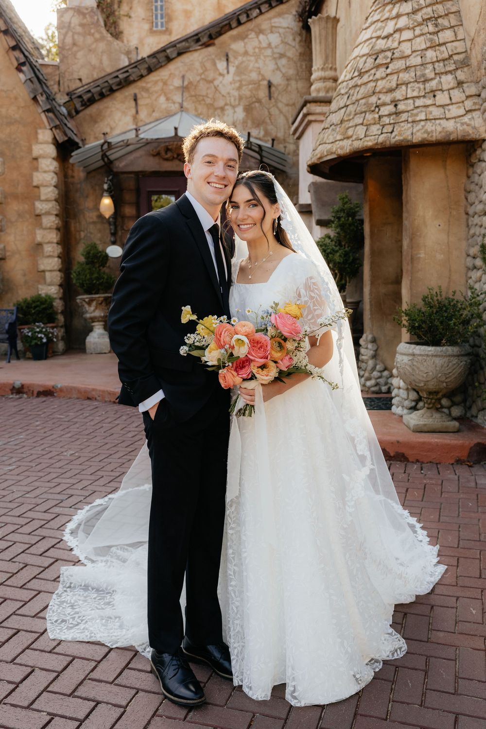 Wedding couple poses in front of rustic stone venue, bride holding colorful spring bouquet.