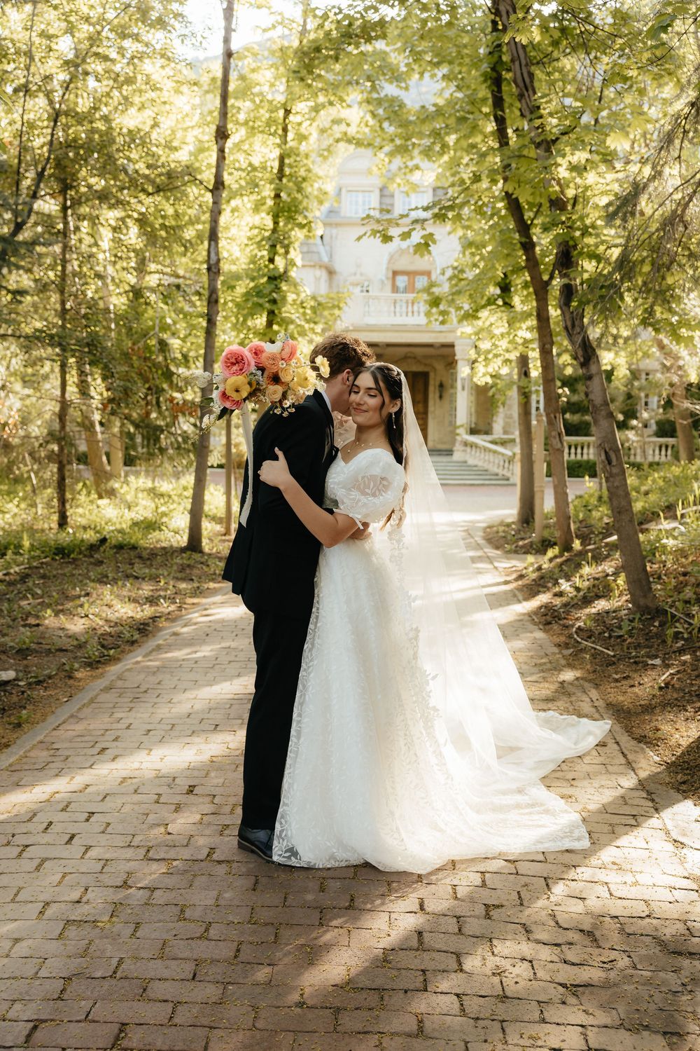 Bride and groom embracing on a tree-lined brick pathway with a grand white manor in the background.