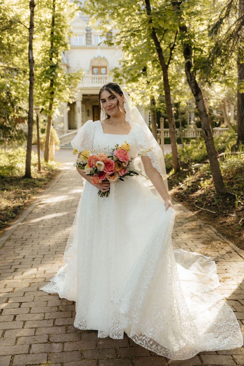 Elegant bride in lace gown holding a colorful bouquet walking along a sunlit tree-lined brick pathway.