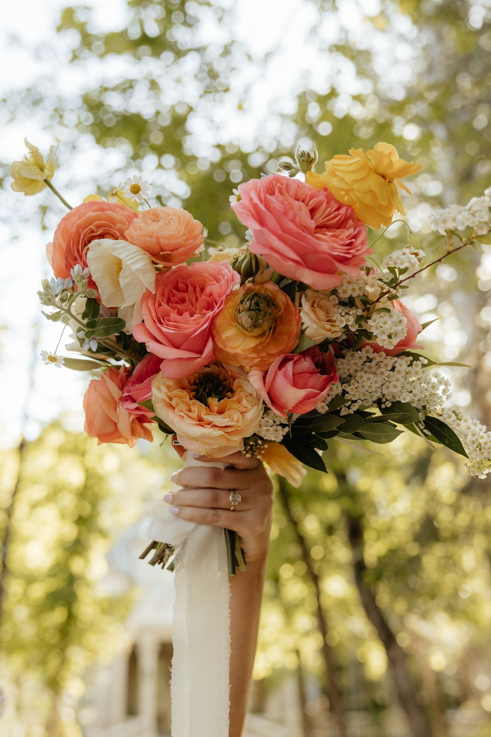 Close-up of bridal bouquet featuring coral roses, ranunculus, and white blooms with ribbon.