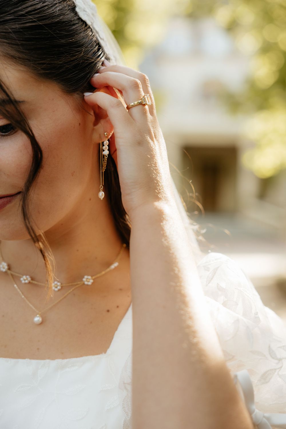 Close-up of bride adjusting gold floral earring, wearing pearl necklace and lace wedding gown.