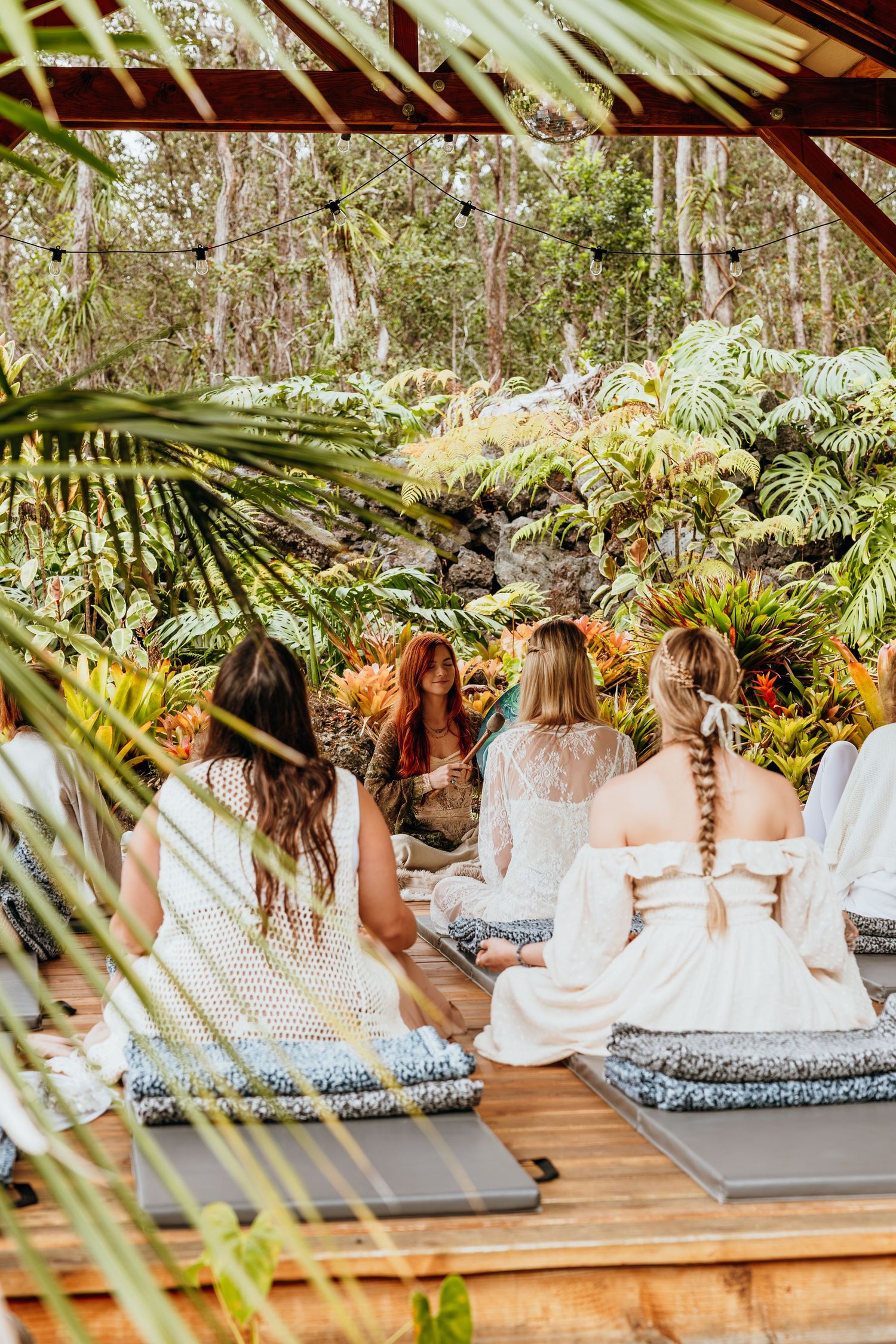 Group in white dresses sitting on cushions in a tropical garden setting.