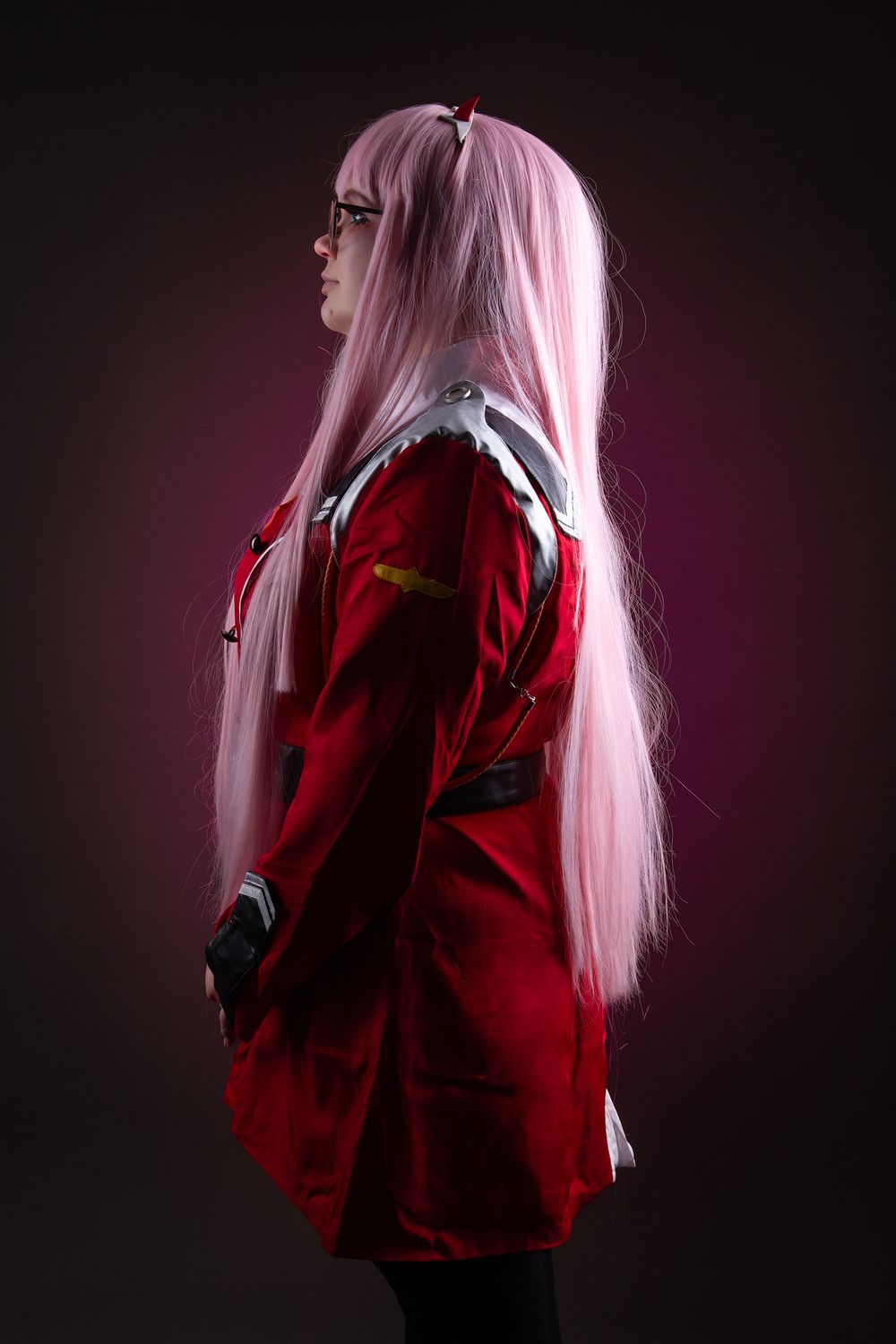 A cosplay figure with long pink hair wearing a red jacket stands in dramatic lighting against a dark background.