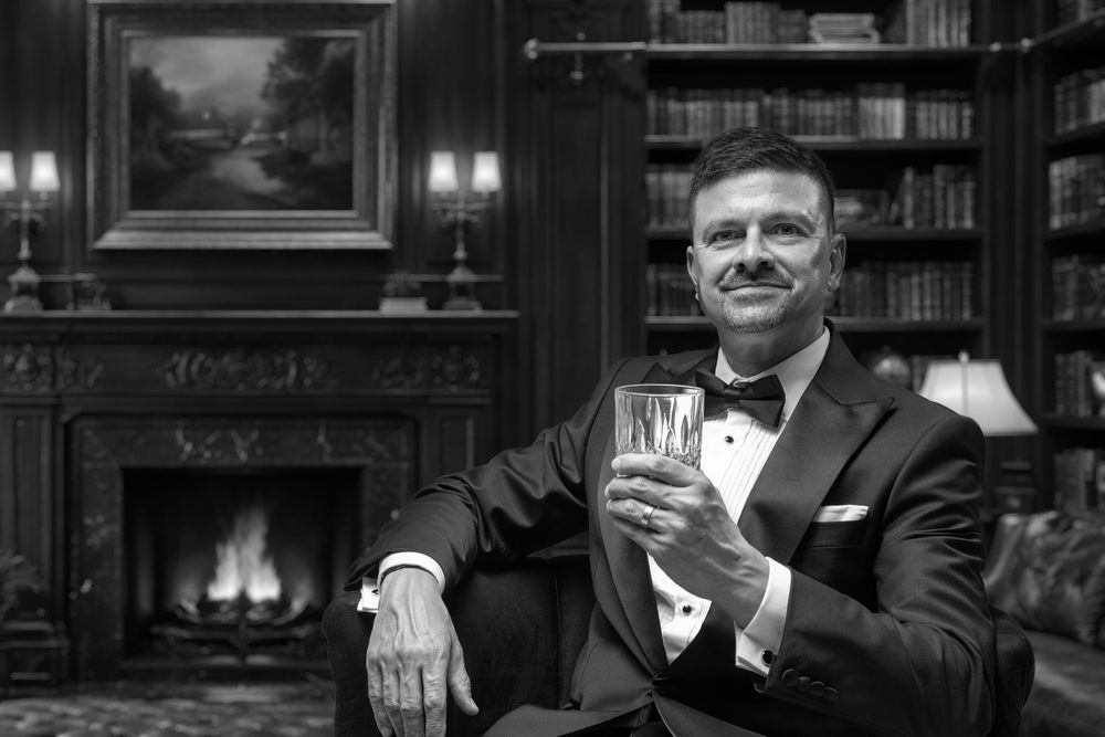 Man in tuxedo holding whiskey glass by fireplace in elegant library, black and white portrait photo.